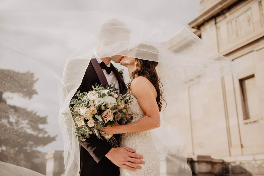 A bride and groom sharing a romantic kiss under a flowing white veil during an intimate At-Home Wedding ceremony.